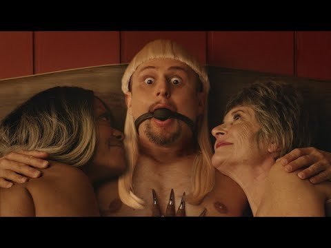Three people: a man with a ball gag and blonde wig is flanked by two women in a close-up shot with a red background.