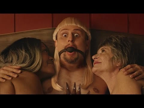 Three people: a man with a ball gag and blonde wig is flanked by two women in a close-up shot with a red background.