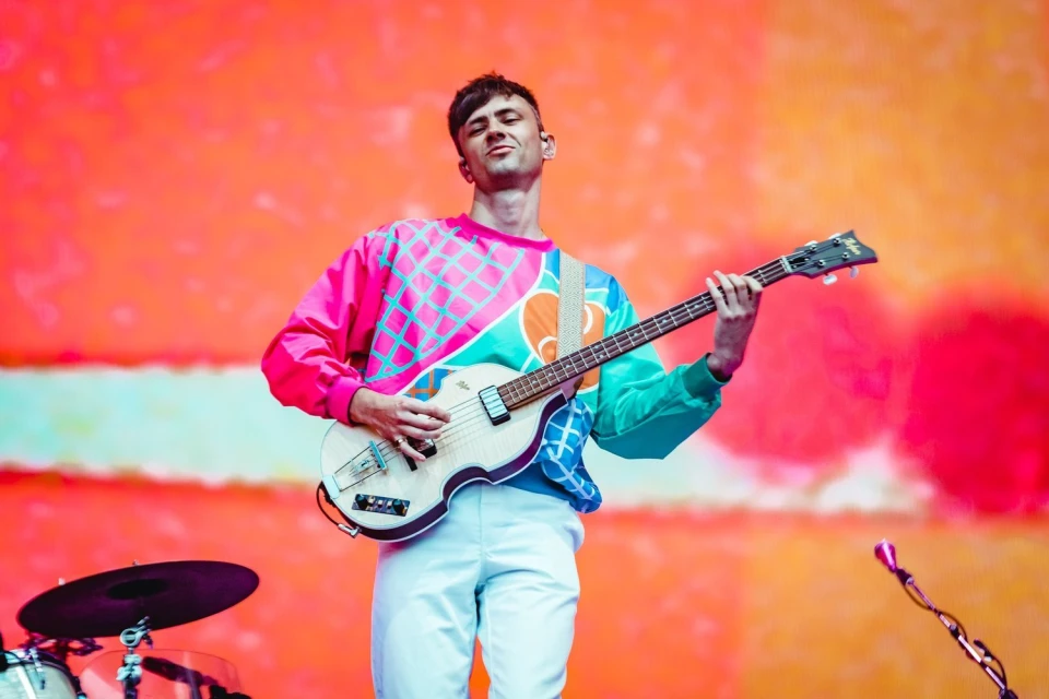 Adult male musician plays white bass guitar in front of an orange background wearing a colorful patterned sweatshirt and white pants.