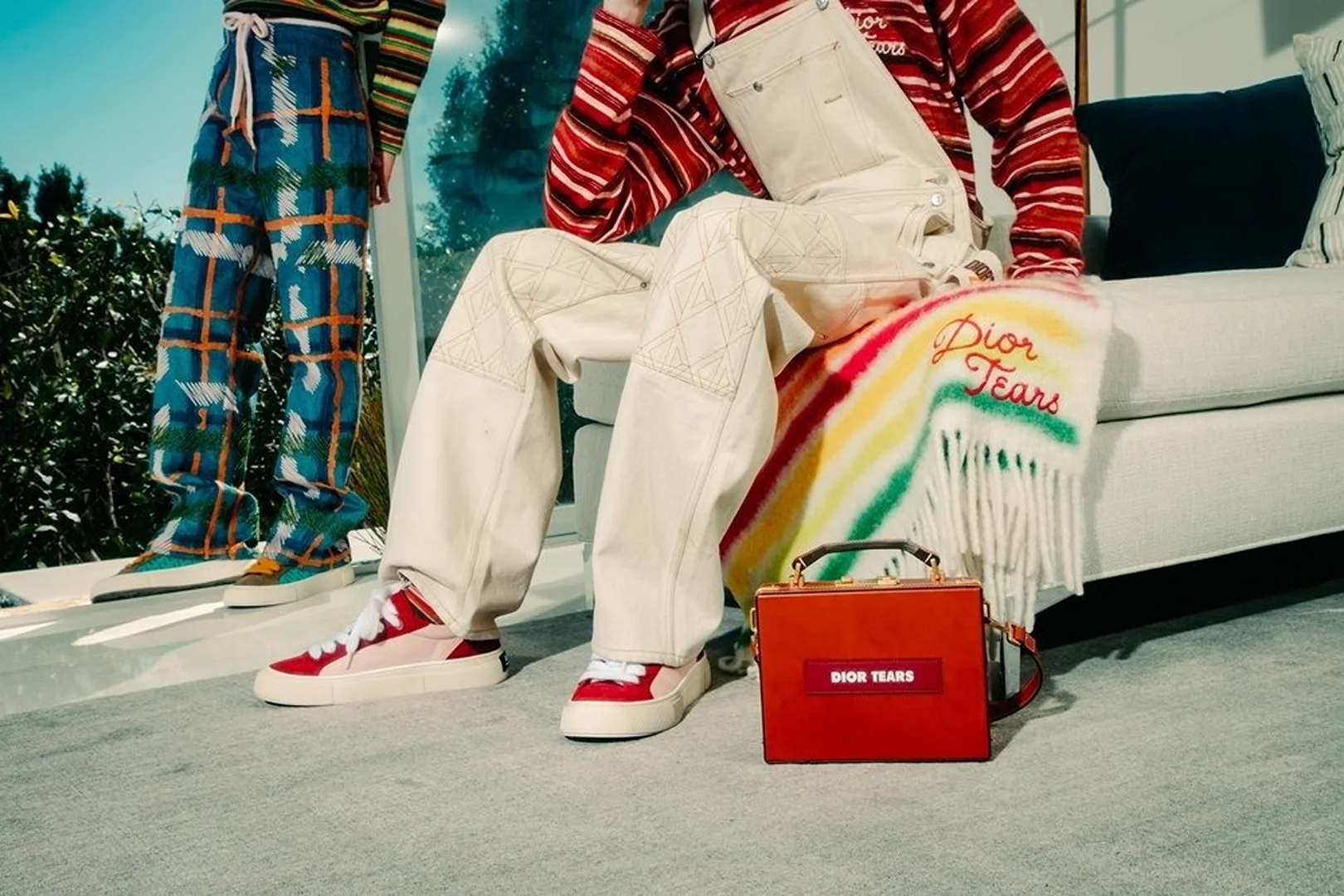 Two fashion models showcase colorful, patterned pants and red sneakers in a casual setting with a Dior Tears accessory bag.