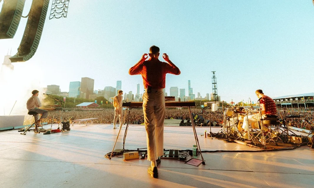 Band performs on stage at outdoor concert with large crowd and city skyline backdrop on a bright day, captured in a wide shot.