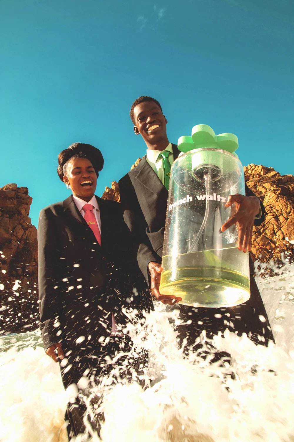 Two people in suits holding a large perfume bottle on a rocky shore with crashing waves, under a bright blue sky, commercial.