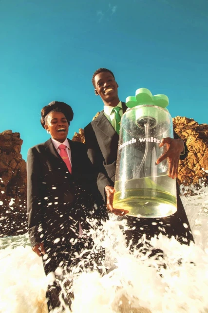 Two people in suits holding a large perfume bottle on a rocky shore with crashing waves, under a bright blue sky, commercial.