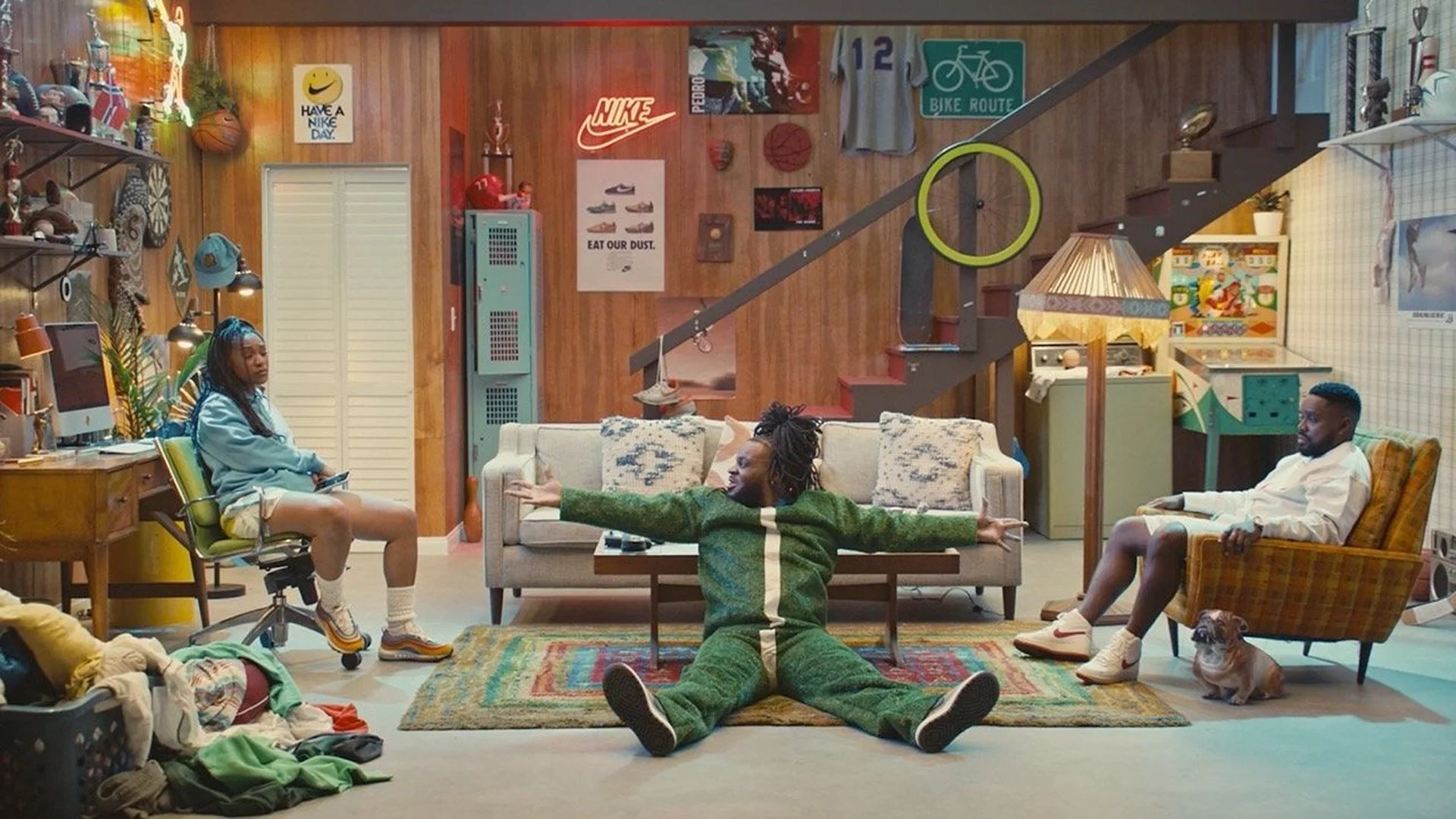Three people in a retro-styled living room: a woman at a desk, a man in a green jumpsuit on the floor, and a man in an armchair.