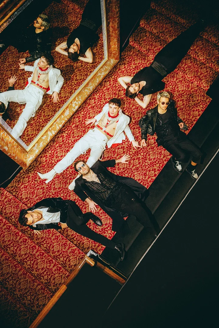 Group of six people in formal wear lying on patterned carpet, shot from overhead angle with high contrast in indoor environment.