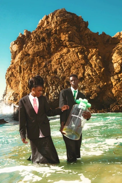Two people in suits stand in shallow water near a rocky cliff carrying a large water bottle in a stylized editorial fashion photograph.