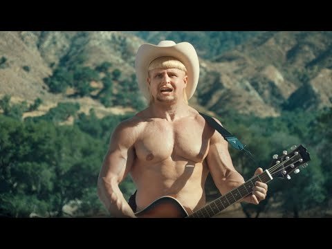 Man with guitar in nature. Muscular man in cowboy hat plays guitar in front of trees and a hill during the daytime.