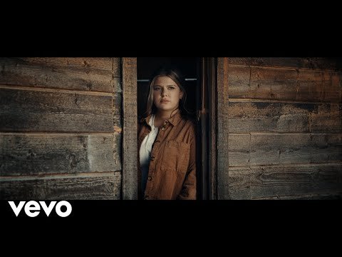 Young woman stands in the doorway of a weathered wood cabin, bathed in soft natural light. She wears a brown jacket and a white top.
