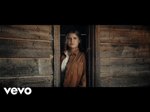 Young woman stands in the doorway of a weathered wood cabin, bathed in soft natural light. She wears a brown jacket and a white top.
