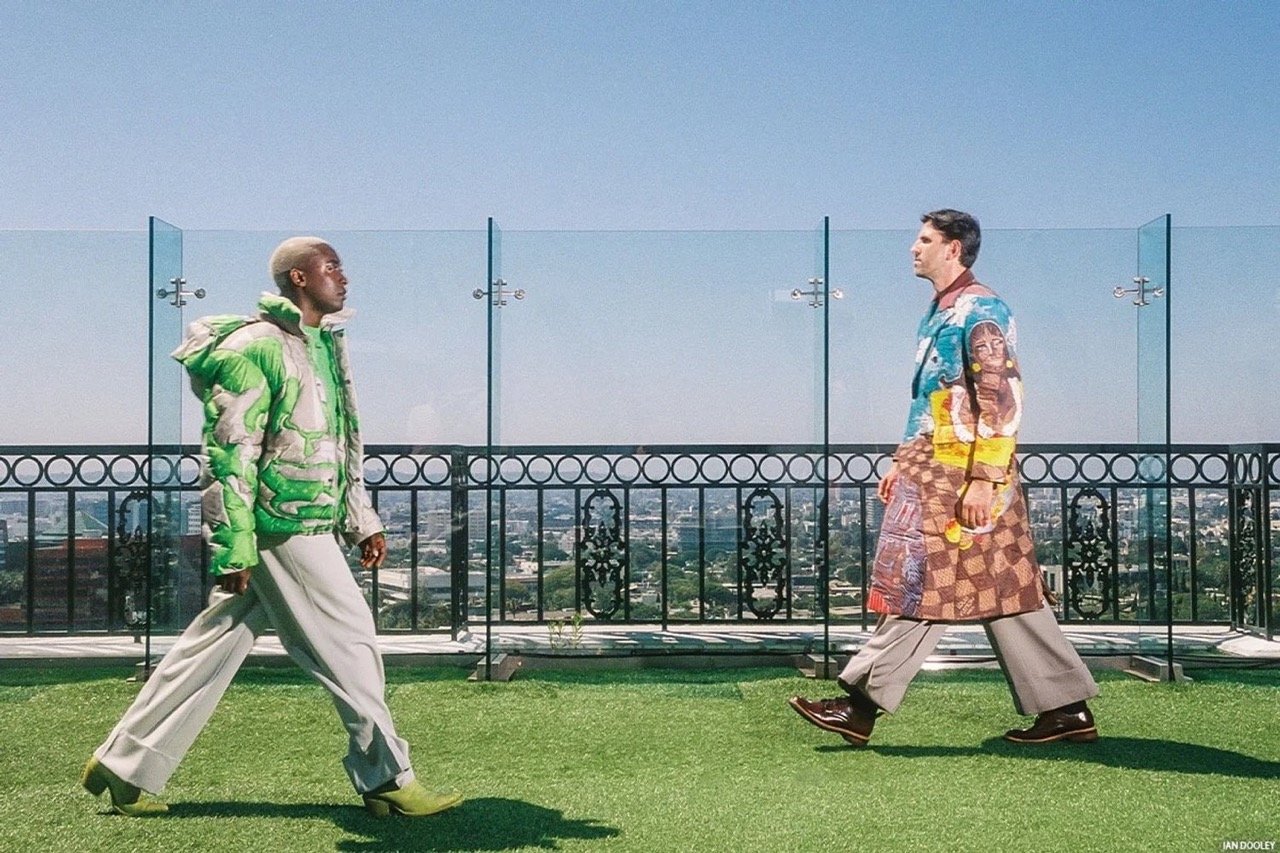 Two fashion models in artistic outerwear walk on a rooftop overlooking a cityscape on a clear, bright day showcasing modern urban fashion.