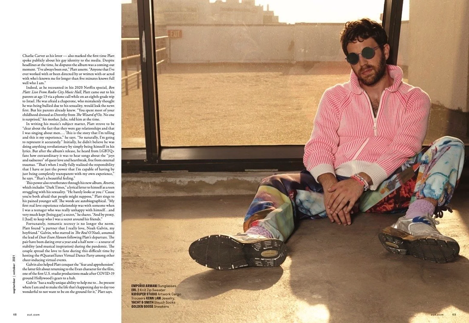 Man sits on concrete floor in striped sweater, patterned pants, round sunglasses. Natural light illuminates an urban, rooftop setting.