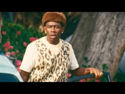 Tyler, The Creator poses in a tan hat and leopard print sweater against a background of green foliage and red flowers.