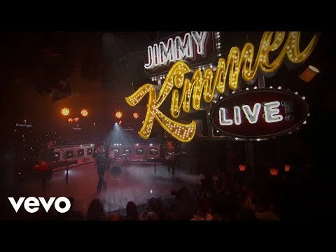A live music performance on the Jimmy Kimmel Live stage with the band, singer, illuminated logo, and a visible audience.