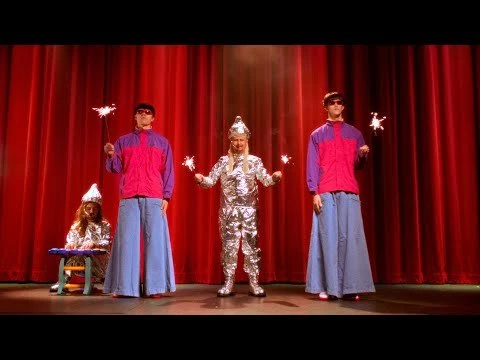 Performance on stage with four people dressed in metallic and colorful costumes, holding sparklers in front of red curtains.