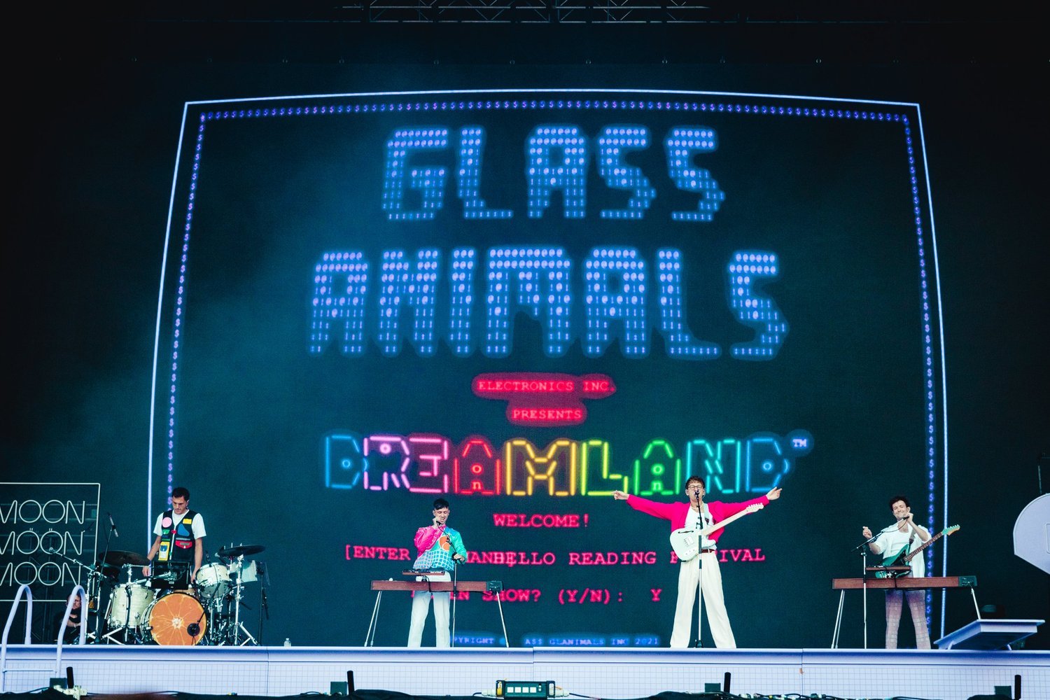 Band Glass Animals performs on stage in front of a large screen displaying the band's name and album title. The group consists of four adult males.