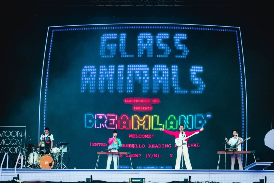 Band Glass Animals performs on stage in front of a large screen displaying the band's name and album title. The group consists of four adult males.