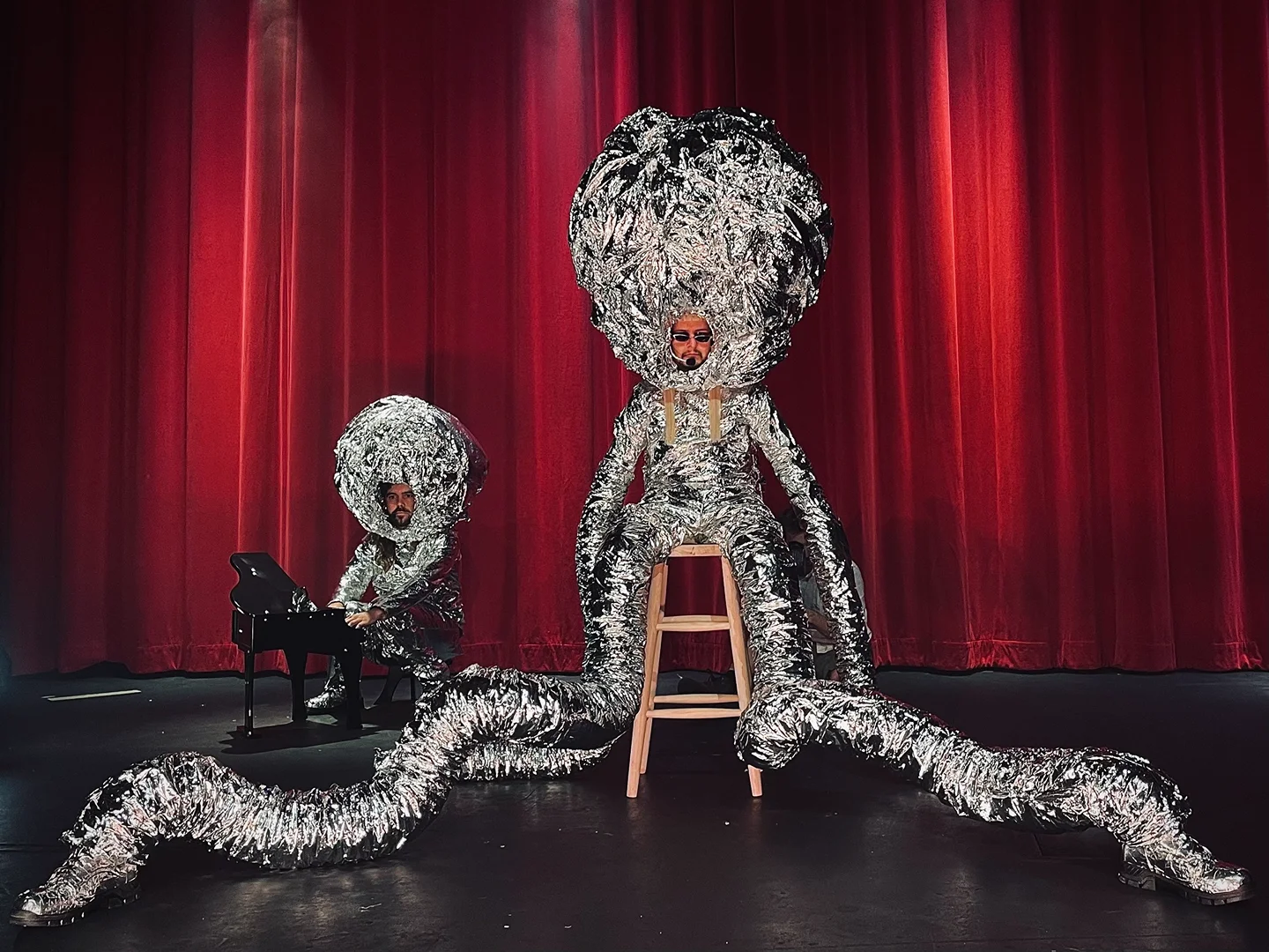 Two people on stage in shiny, silver costumes performing in front of a red curtain with one playing a piano and the other sitting.