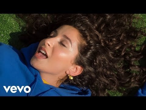 Close-up of a woman with curly hair lying on green grass, wearing a blue sweatshirt and yellow shirt. She has gold earrings.
