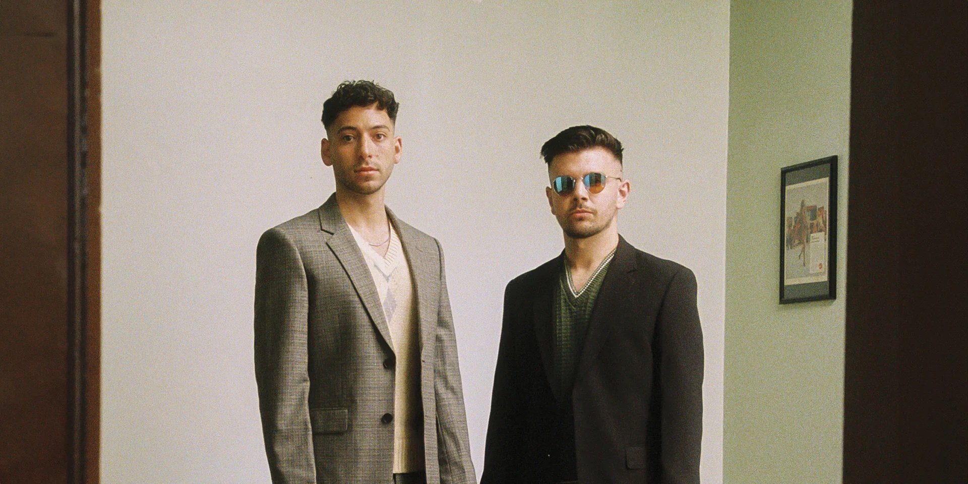 Two adult men pose indoors wearing suit jackets and knit tops, with one wearing sunglasses. A picture frame hangs on the wall.