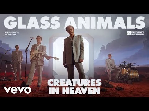 Music video still features members of Glass Animals in suits performing; stage with a desert backdrop and musical instruments visible.