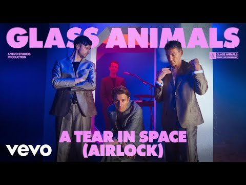 Four members of Glass Animals stand in suits in a music video scene with blue and pink lighting, promoting their song, 'A Tear in Space'.