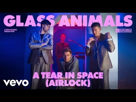 Four members of Glass Animals stand in suits in a music video scene with blue and pink lighting, promoting their song, 'A Tear in Space'.