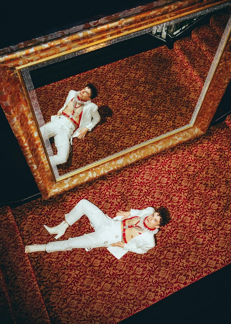 A young adult lies on an ornate red and gold patterned carpet in a white suit reflected in a gold-framed mirror from an overhead angle.