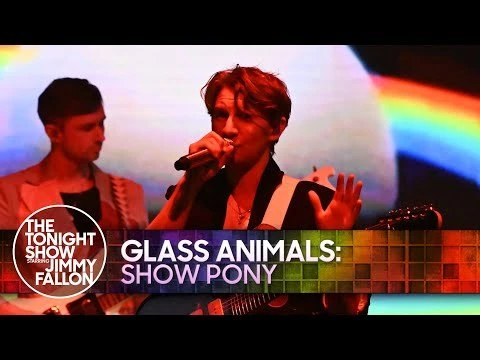 Two male musicians perform on stage with guitars and a microphone; rainbow lighting background and TV show logo in the lower left.