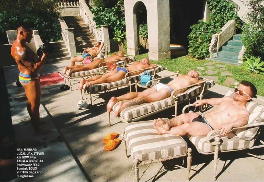 A muscular man lifts weights in a rainbow swimsuit, with other men in swimwear lounging on chairs and tanning outdoors in bright sunlight.