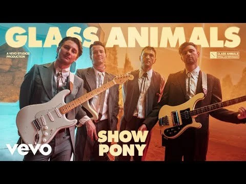 Four musicians stand with guitars and bass guitars in an indoor music video setting, against a stylized backdrop, promoting Glass Animals and their song.