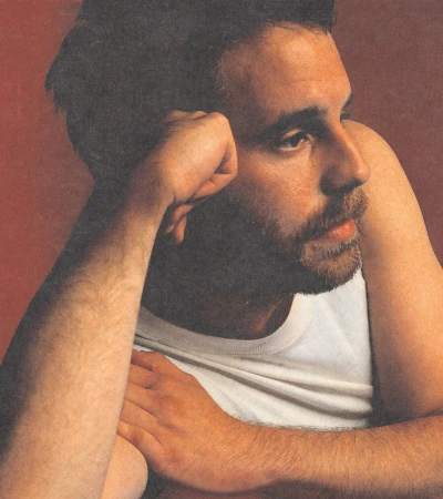 Close-up portrait of a man with a beard wearing a white tank top against a dark reddish-brown background, styled by Kenn Law.