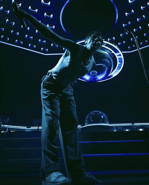 Singer performing on stage with microphone, wearing a white tank top and dark pants with spotlight lighting and stage equipment.