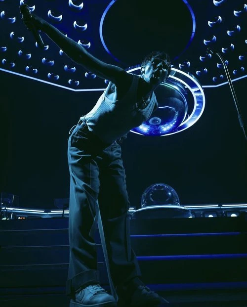 Singer performing on stage with microphone, wearing a white tank top and dark pants with spotlight lighting and stage equipment.