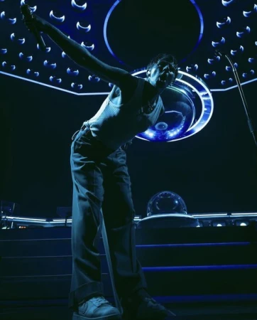 Singer performing on stage with microphone, wearing a white tank top and dark pants with spotlight lighting and stage equipment.