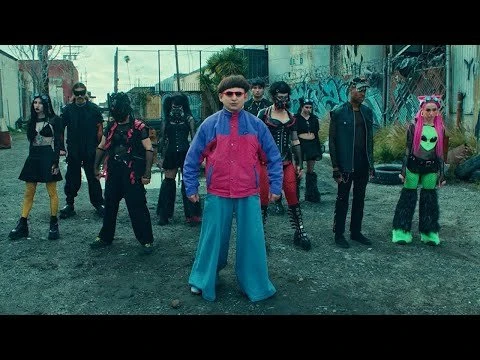 A group of people in alternative fashion pose in an urban alleyway, featuring brightly colored outfits, masks, and bold accessories.