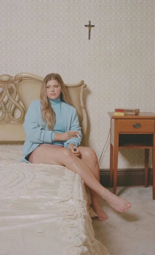 Woman with long blonde hair sits on a bed in a blue sweater smoking a cigarette with a nightstand and crucifix in the background.