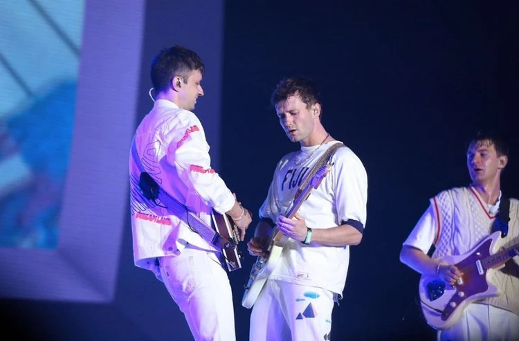 Three young adult males are shown performing live on stage playing electric guitars, bathed in soft, cool lighting, showcasing their musical talent.