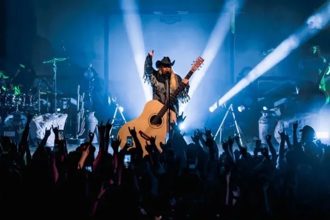 A musician with long blonde hair, wearing a cowboy hat and a fringed jacket, plays an acoustic guitar on stage in front of a crowd.