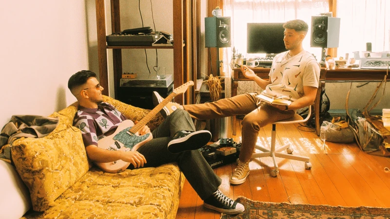 Two men in a relaxed indoor music studio setting, one playing guitar on a mustard-colored couch, the other seated with a notebook.