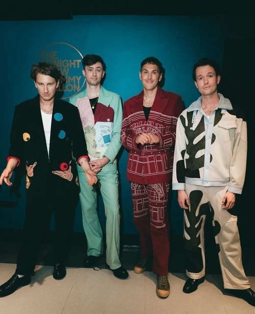 Four young adult men stand in front of a teal wall with 'The Tonight Show Starring Jimmy Fallon' logo, wearing fashion-forward suits.