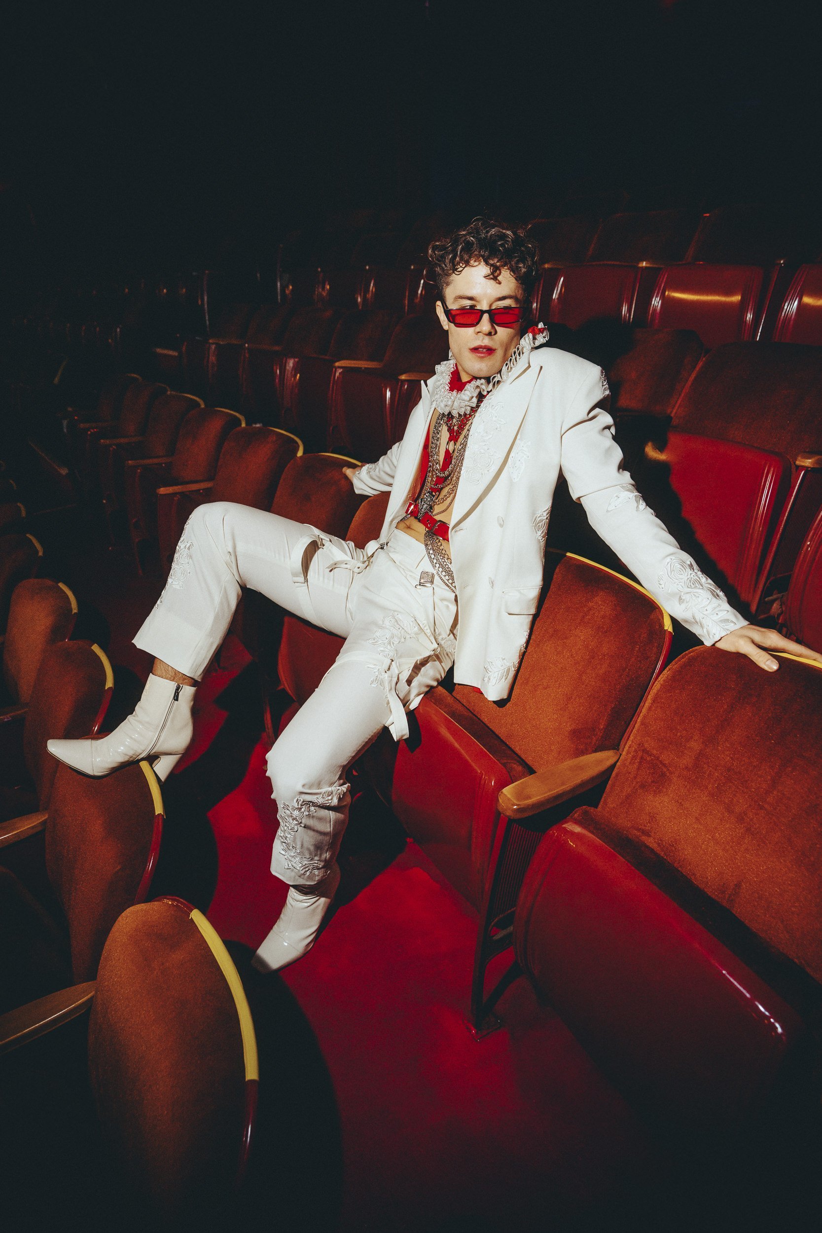 Stylish man in white suit poses in theatre seating, with red sunglasses and boots, showcasing fashion and individual expression.