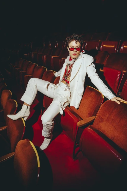 Stylish man in white suit poses in theatre seating, with red sunglasses and boots, showcasing fashion and individual expression.