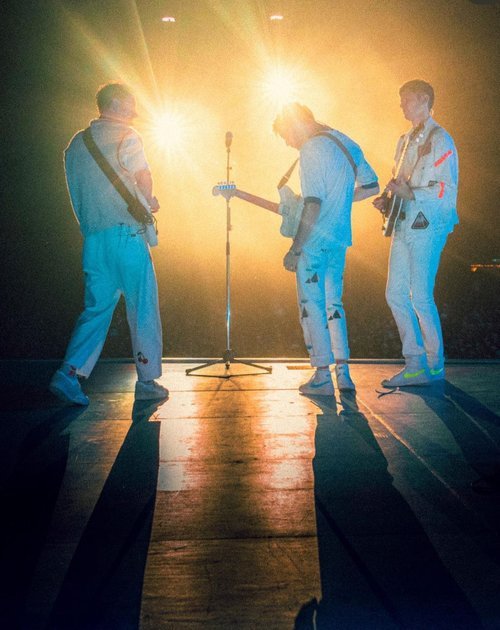 Band of three men performing on stage with guitars in matching white outfits backlit by bright yellow lights, creating long shadows.
