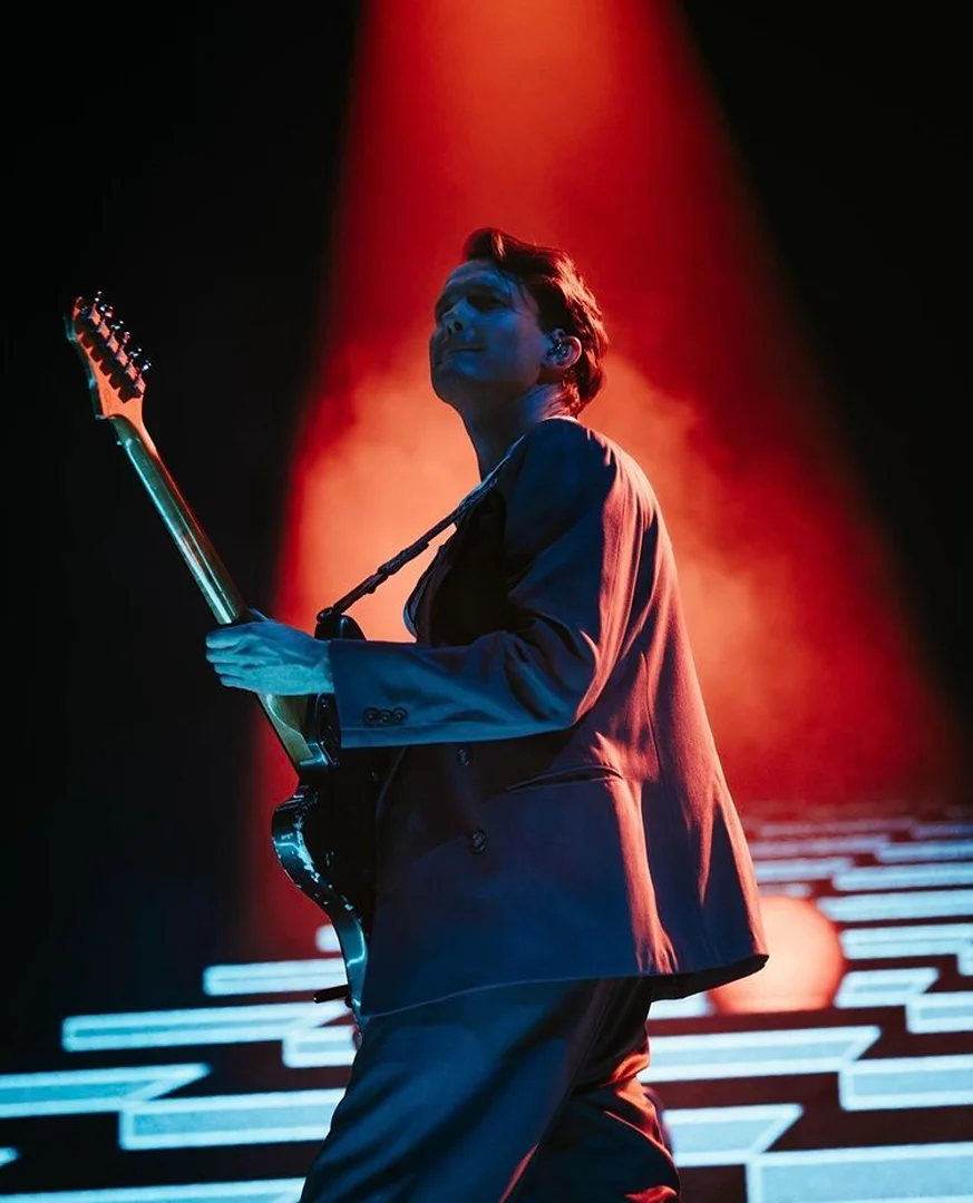 Male musician wearing a suit plays electric guitar on stage, lit by bright red and blue spotlights during a concert performance.