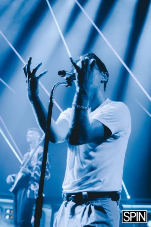 Singer performs on stage with microphone in hand under blue spotlights, guitarist in background, concert photography, music performance.