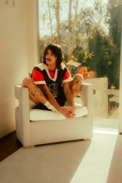 Young adult with mustache and tattoos sits barefoot in a white armchair, relaxing in a brightly lit room with floor-to-ceiling windows.