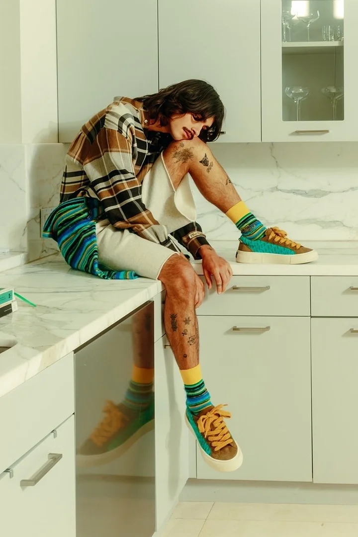 Young adult sitting on a kitchen counter wearing a plaid shirt, shorts, socks, and sneakers in a casual and fashionable pose.