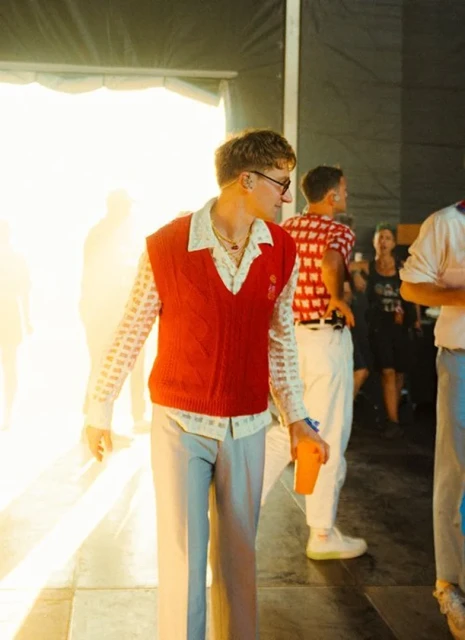 A young adult male with blond hair stands indoors wearing a red knit vest, patterned shirt, and light trousers at an indoor venue.