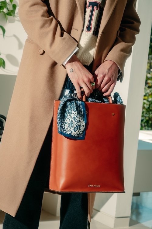 A fashionable person carrying a tan leather tote bag with a floral scarf tied to the handle, styled with a camel coat and patterned sweater.