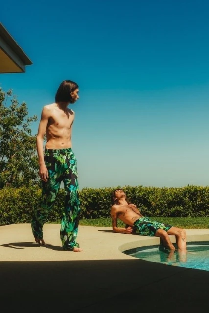 Two shirtless men in tropical-print bottoms relax poolside in bright daylight under a clear blue sky surrounded by green foliage.